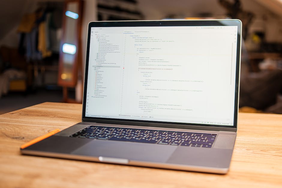 A modern laptop placed on a wooden table displaying lines of code or a programming interface, with a blurred background showing a cozy indoor space with warm lighting, shelves, and possibly household